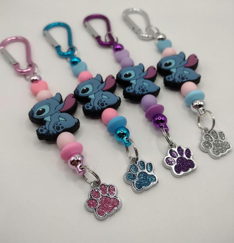 May include: Keychains featuring a blue cartoon character with large ears and a paw print charm. The keychains have colorful beads in pink, blue, and purple, and silver-tone carabiner clips.