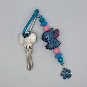 Könnte beinhalten: Ein blauer Metall-Karabiner-Schlüsselanhänger mit einem silbernen Schlüssel, einer blau-rosa Perlenschnur, einem blauen Zeichentrickfiguren-Anhänger und einem baumelnden, blau glitzernden Pfotenabdruck-Anhänger. Der Schlüssel hat die Buchstaben HD.