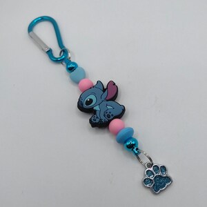 May include: A blue and pink beaded keychain featuring a cartoon character and a paw print charm. The keychain has a blue carabiner clip, pink and blue beads, and a blue cartoon character. The charm is a blue paw print.