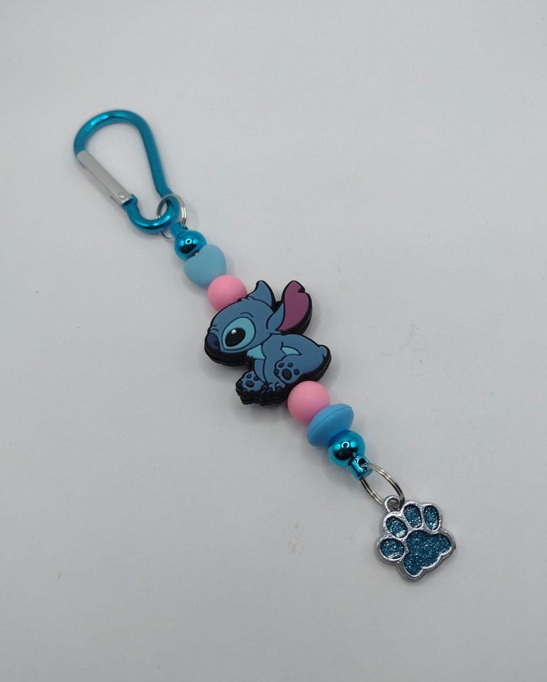 May include: A blue and pink beaded keychain featuring a cartoon character and a paw print charm. The keychain has a blue carabiner clip, pink and blue beads, and a blue cartoon character. The charm is a blue paw print.
