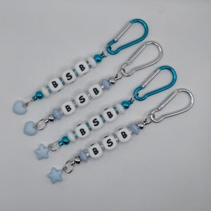 Puede incluir: Cuatro llaveros con cuentas blancas que muestran las letras negras B, S y B. Cada llavero incluye cuentas decorativas azules y plateadas, un dije de corazón o estrella azul claro en el extremo y un mosquetón metálico en color azul o plata.