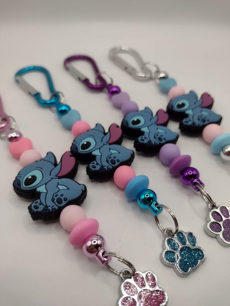 May include: Several keychains featuring a blue cartoon character with large ears and a paw print charm. The keychains have a variety of colorful beads in pink, blue, and purple. Each keychain has a silver clasp.