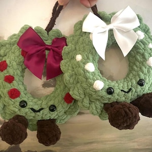 May include: Two handmade, green crochet wreaths with smiling faces and brown feet. One wreath has a burgundy bow, the other a white bow. Red and white accents dot the wreaths, held by a brown loop.