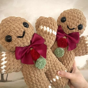 May include: Two handmade plush gingerbread men with brown bodies, black button eyes, and smiling faces. Each has a burgundy bow, a red pom-pom, and a green pom-pom. White stitching outlines the arms and legs.