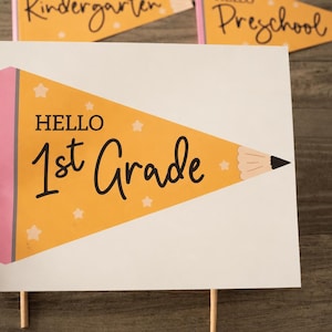 May include: A rectangular sign featuring a yellow pencil-shaped pennant design with a pink eraser and black text that reads "HELLO 1st Grade" with small white star accents. The sign is mounted on wooden sticks, with other similar signs visible.