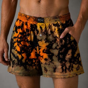May include: Boxer shorts featuring a black and orange tie-dye pattern with an elastic waistband. The front includes a central button closure and a black label with the white text KOMPEDAN and the size XL printed on the centre of the waistband.