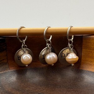 Puede incluir: Tres marcadores de punto de plata con cuentas de perlas y letras grabadas "B", "I" y "D".