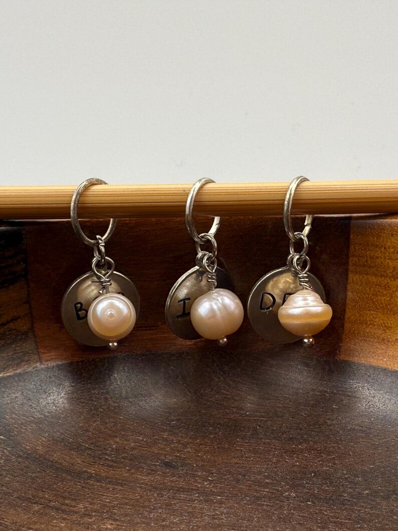 Puede incluir: Tres marcadores de punto de plata con cuentas de perlas y letras grabadas "B", "I" y "D".