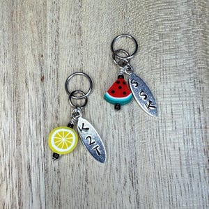May include: Two silver knitting stitch markers with fruit charms. One has a yellow lemon slice, the other a red watermelon slice. Each has a silver tag with the letters "K2T" and "SSK".