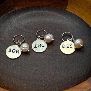 Puede incluir: Tres abalorios de plata con detalles de perlas. Cada abalorio tiene una abreviatura de mes diferente: BOR, INC y DEC.