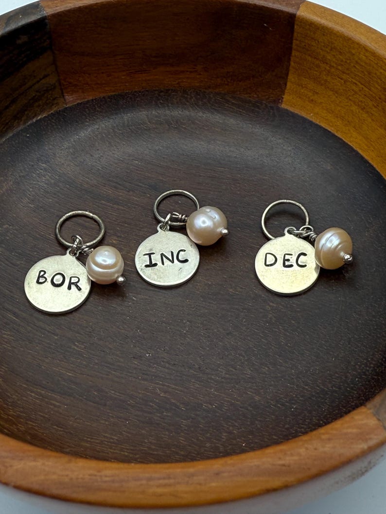 Puede incluir: Tres abalorios de plata con detalles de perlas. Cada abalorio tiene una abreviatura de mes diferente: BOR, INC y DEC.