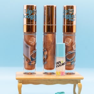 May include: Three gold lip gloss tubes with a peace sign and the text "Lip Doogan!" on the label. The tubes are on a wooden table with gold coins and blue glass gems scattered around. The text "Money Honey" is at the top of the image.