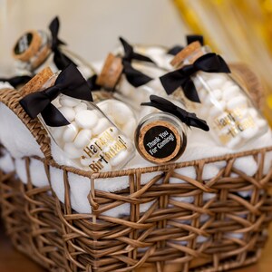May include: A wicker basket filled with glass jars containing white candies. Each jar is adorned with a black bow and a cork stopper. Some jars have gold text that reads "Happy Birthday!" and "Thank You For Coming!"