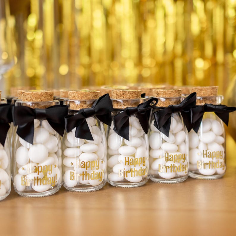 May include: Clear glass bottles filled with white candies, topped with corks and black bows. The bottles have gold text that reads "Happy Birthday". The background features gold streamers.