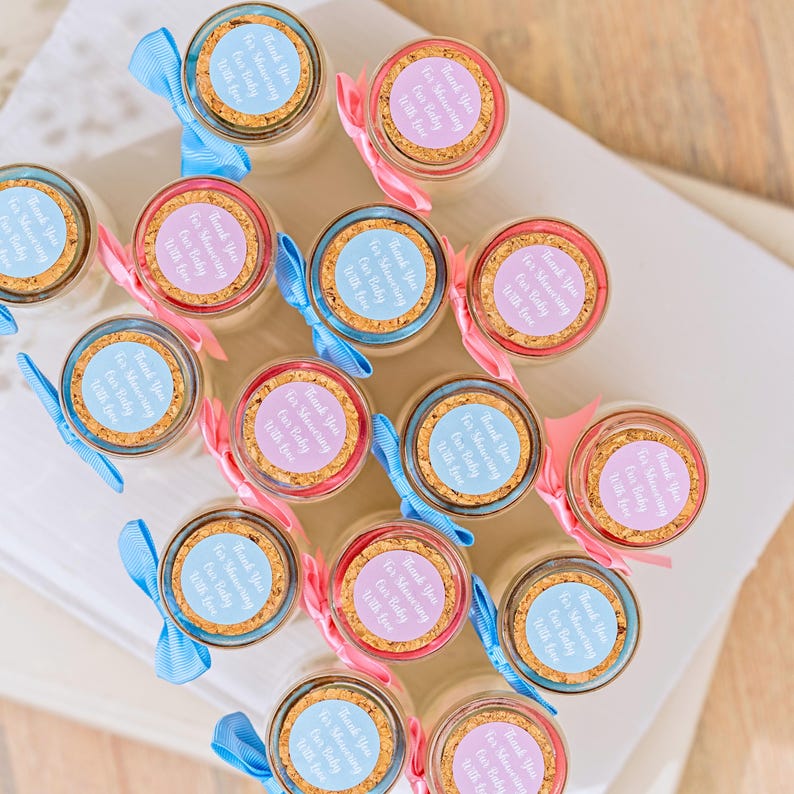 K&ouml;nnte beinhalten: Kleine Gl&auml;ser mit Korkdeckeln, jeweils mit einem runden Etikett und einem Band versehen. Die Etiketten lauten "Thank You For Showering Our Baby With Love." B&auml;nder sind hellblau oder rosa. Ein Geschenk f&uuml;r eine Babyparty.