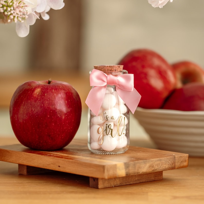K&ouml;nnte beinhalten: Ein kleines Glas mit wei&szlig;en Bonbons, einer rosa Schleife und einem Korken. Auf dem Glas steht in Gold "it's a girl!". Ein gro&szlig;er roter Apfel liegt auf einem Holztablett neben dem Glas. Weitere rote &Auml;pfel sind im Hintergrund.