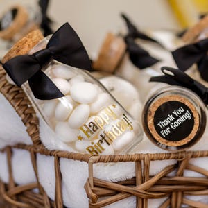 May include: Clear glass jars filled with white candies, adorned with black bows and cork stoppers. One jar has gold text reading "Happy Birthday!" and another has a label that says "Thank You For Coming!" The jars are in a woven basket.