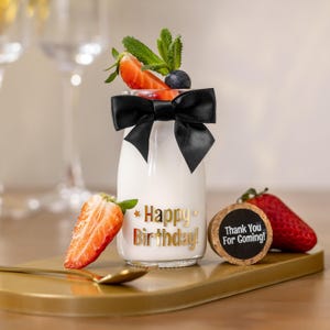 May include: A small glass bottle filled with a white liquid, adorned with a black bow and topped with strawberries, blueberries, and mint. The bottle has gold text that reads "Happy Birthday!" and is on a gold tray.