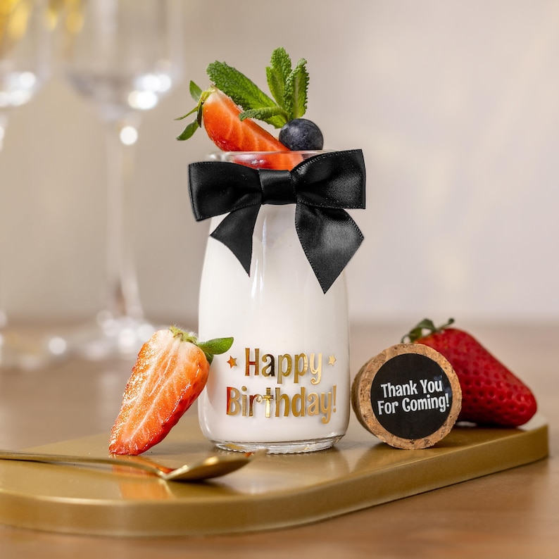 May include: A small glass bottle filled with a white liquid, adorned with a black bow and topped with strawberries, blueberries, and mint. The bottle has gold text that reads "Happy Birthday!" and is on a gold tray.