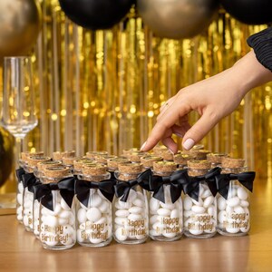 May include: Small glass bottles filled with white candies, each with a cork stopper and a black ribbon. The bottles have "Happy Birthday!" printed in gold. The background features gold and black balloons and a gold fringe.