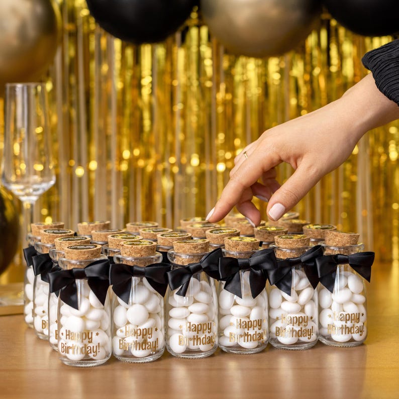 May include: Small glass bottles filled with white candies, each with a cork stopper and a black ribbon. The bottles have "Happy Birthday!" printed in gold. The background features gold and black balloons and a gold fringe.