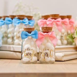K&ouml;nnte beinhalten: Gl&auml;ser gef&uuml;llt mit wei&szlig;en Bonbons, mit Korken und Schleifen versehen. Ein Glas hat eine blaue Schleife und sagt "it's a boy!" in goldener Schrift. Ein anderes hat eine rosa Schleife und sagt "it's a girl!"