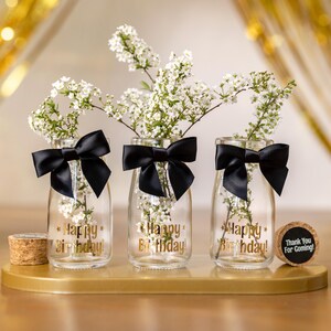 May include: Three clear glass vases with white flowers, black bows, and gold text that reads "Happy Birthday!". A cork with "Thank You For Coming!" is also present. The vases are on a gold tray.