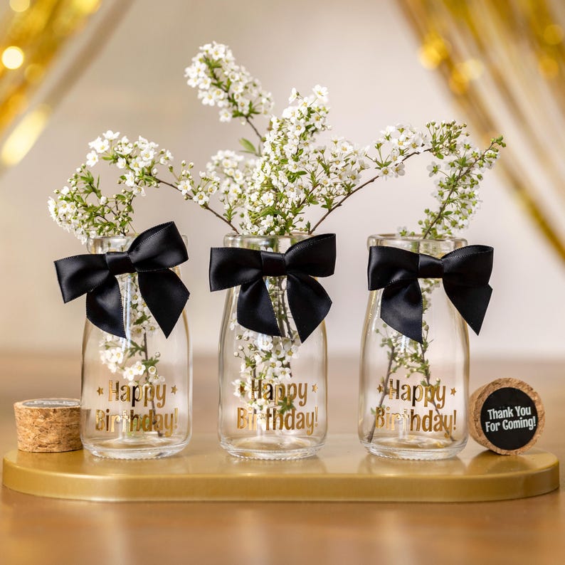 May include: Three clear glass vases with white flowers, black bows, and gold text that reads "Happy Birthday!". A cork with "Thank You For Coming!" is also present. The vases are on a gold tray.