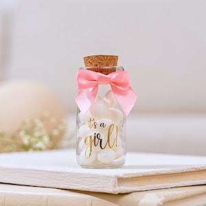 K&ouml;nnte beinhalten: Ein kleines Glasgef&auml;&szlig; gef&uuml;llt mit wei&szlig;en S&uuml;&szlig;igkeiten, gekr&ouml;nt mit einem Korken und einer rosa Satinschleife. Das Glas hat goldfarbene Schrift, die "It's a girl!" lautet. Ein charmantes Geschenk f&uuml;r eine Babyparty.