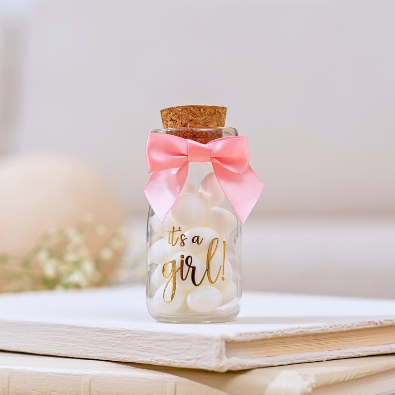 K&ouml;nnte beinhalten: Ein kleines Glasgef&auml;&szlig; gef&uuml;llt mit wei&szlig;en S&uuml;&szlig;igkeiten, gekr&ouml;nt mit einem Korken und einer rosa Satinschleife. Das Glas hat goldfarbene Schrift, die "It's a girl!" lautet. Ein charmantes Geschenk f&uuml;r eine Babyparty.