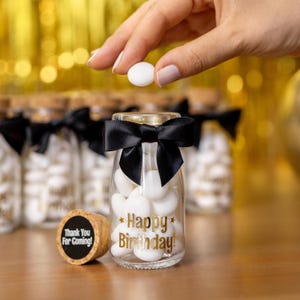 May include: A clear glass jar filled with white candies, tied with a black bow, and the words "Happy Birthday!" in gold. A cork with "Thank You For Coming!" is next to the jar. A hand is holding two white candies above the jar.