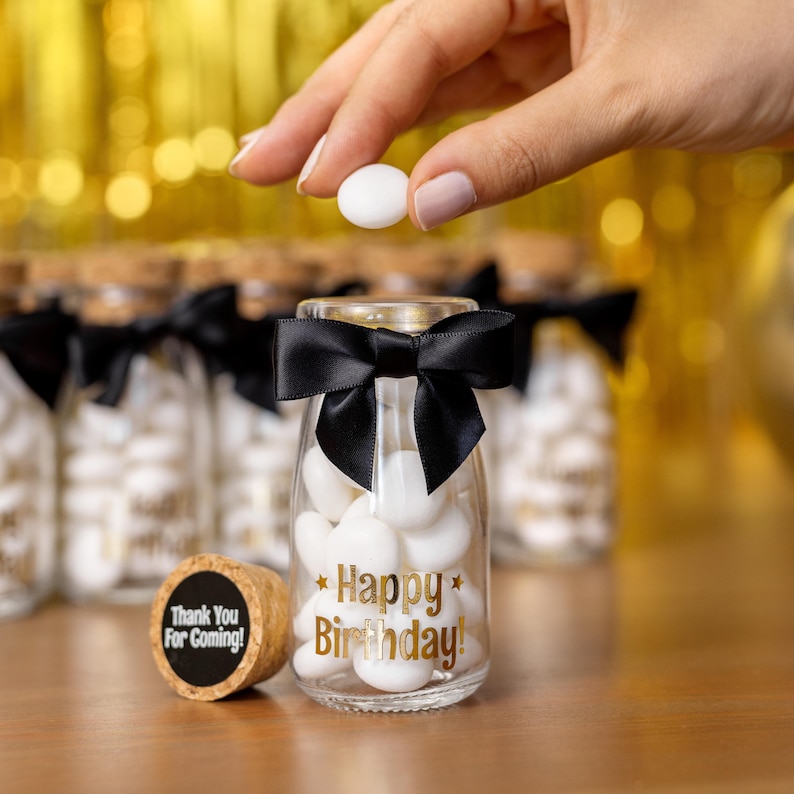 May include: A clear glass jar filled with white candies, tied with a black bow, and the words "Happy Birthday!" in gold. A cork with "Thank You For Coming!" is next to the jar. A hand is holding two white candies above the jar.