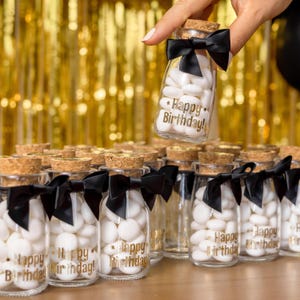 May include: Small glass bottles filled with white candies, each with a cork stopper and a black bow. The bottles are labeled with the text "Happy Birthday!" in gold lettering. A hand holds one bottle above the others.