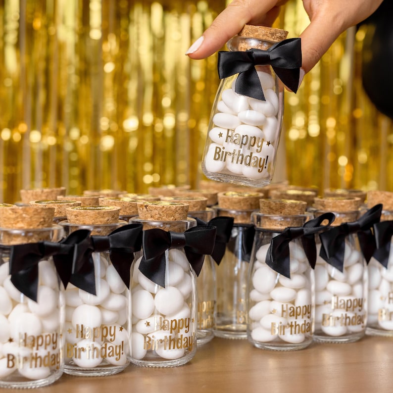 May include: Small glass bottles filled with white candies, each with a cork stopper and a black bow. The bottles are labeled with the text "Happy Birthday!" in gold lettering. A hand holds one bottle above the others.