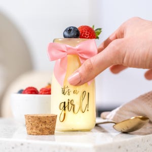K&ouml;nnte beinhalten: Kleine Glasflasche gef&uuml;llt mit einer gelben Fl&uuml;ssigkeit, verziert mit einer rosa Schleife und belegt mit einer Blaubeere und einer Erdbeere. Die Flasche tr&auml;gt den Text "it's a girl!" in goldener Schrift. Ein Korken und ein L&ouml;ffel sind ebenfalls sichtbar.