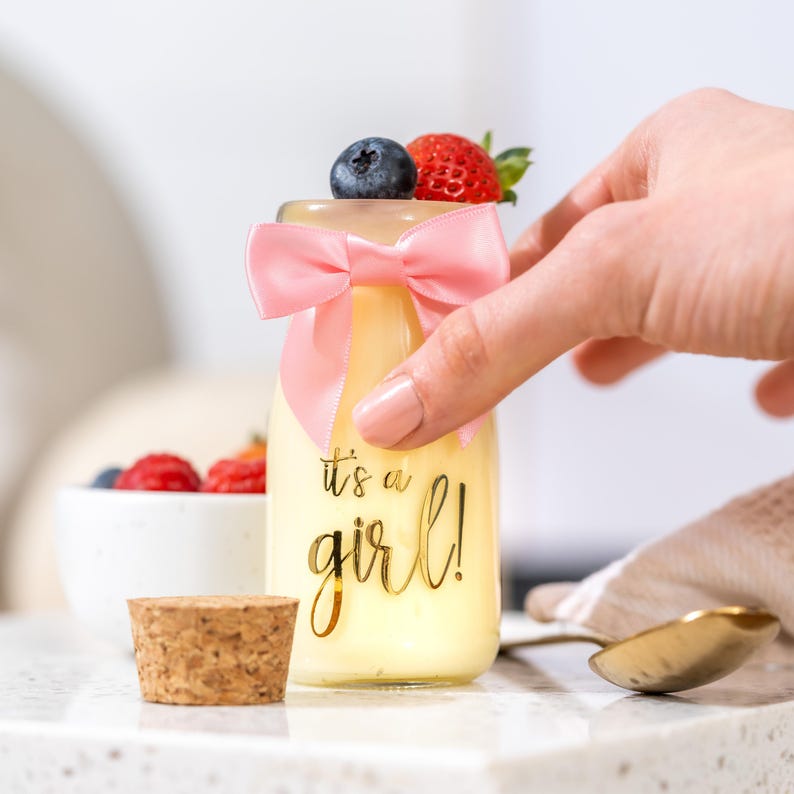 K&ouml;nnte beinhalten: Kleine Glasflasche gef&uuml;llt mit einer gelben Fl&uuml;ssigkeit, verziert mit einer rosa Schleife und belegt mit einer Blaubeere und einer Erdbeere. Die Flasche tr&auml;gt den Text "it's a girl!" in goldener Schrift. Ein Korken und ein L&ouml;ffel sind ebenfalls sichtbar.