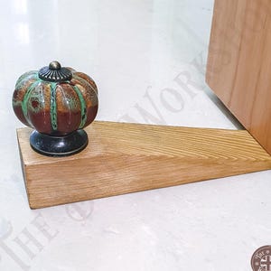 May include: A wooden doorstop featuring a wedge-shaped base and a decorative ceramic knob. The knob has a pumpkin-like shape with a glossy finish in shades of brown and green. The text The Brit Workshop is visible on the image.