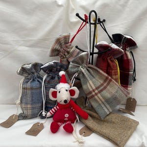 May include: A selection of tartan fabric drawstring bags, alongside a small, plush mouse dressed in a Santa hat and red clothing. The bags feature patterns in blue, red, and brown, with attached tags.