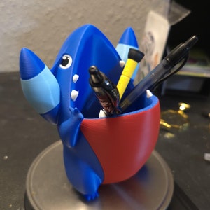 May include: Blue and red 3D printed Pokemon character with a large open mouth, holding pens and pencils. The character is sitting on a silver scale.