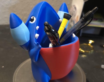 May include: Blue and red 3D printed Pokemon character with a large open mouth, holding pens and pencils. The character is sitting on a silver scale.