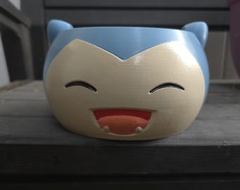 May include: A round ceramic planter featuring the face of a Snorlax character. The planter is light blue with a cream-coloured face, black closed eyes, and an open mouth showing a red tongue and two small white teeth, with two small ears on top.