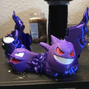 Pode incluir: Figuras de Pokémon Gengar, Haunter e Gastly impressas em 3D na cor roxa. As figuras estão dispostas em círculo com chamas atrás delas.