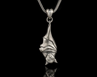 May include: A silver necklace with a detailed bat pendant hanging from it. The bat is upside down with its wings spread out. The necklace is on a black background.