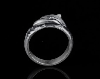 May include: A silver ring with a dolphin design. The ring is on a black background and is reflecting light.