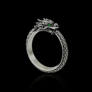May include: A silver dragon ring with a detailed dragon head and green gemstone eyes. The ring's band has a scale pattern. The ring is set against a black background. A unique piece of jewellery.