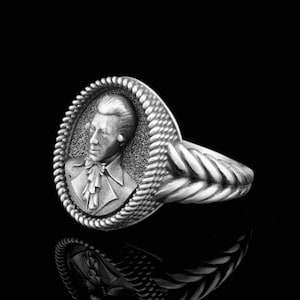 Può includere: Un anello d'argento con un ritratto dettagliato di un uomo in un cammeo. La fascia dell'anello è intrecciata.