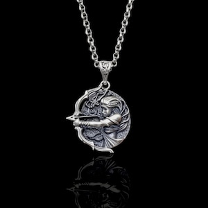May include: A silver pendant necklace featuring a detailed circular medallion. The medallion depicts a figure with a bow and arrow, surrounded by ornate floral designs. The necklace has a delicate chain and a decorative clasp.