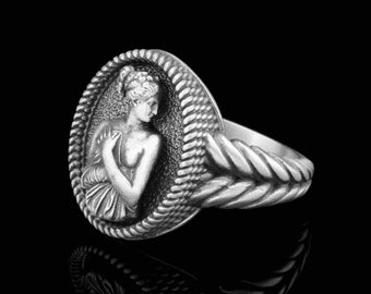 May include: A silver ring with a detailed design featuring a woman's profile in a circular setting. The band of the ring is braided.