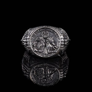 Op de afbeelding: Een zilveren ring met een ingewikkeld ontwerp met een figuur die een zwaard vasthoudt. De ring is geplaatst tegen een zwarte achtergrond.