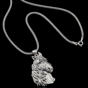 Puede incluir: Un collar plateado con un colgante detallado de cabeza de caballo. El colgante muestra el perfil del caballo con una melena y rasgos faciales detallados. La cadena tiene un dise&ntilde;o tejido y el cierre es visible. El fondo es negro.
